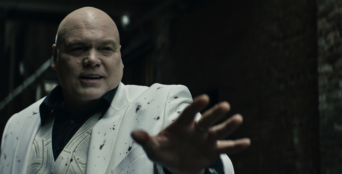 In an image from Marvel Studios’ Echo, Wilson Fisk (Vincent D’Onofrio) is wearing his iconic white suit with black button-up shirt and is gesturing at something or someone off camera to the right. He’s in a darkened room.