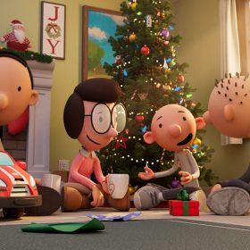 The family from Diary of a Wimpy Kid Christmas: Cabin Fever all sit on the floor in the house in front of a decorated Christmas tree including (from left to right) Manny, Frank, Susan, Greg, and Rodrick. There are stockings hung above the fireplace in the background and the family is smiling at each other.