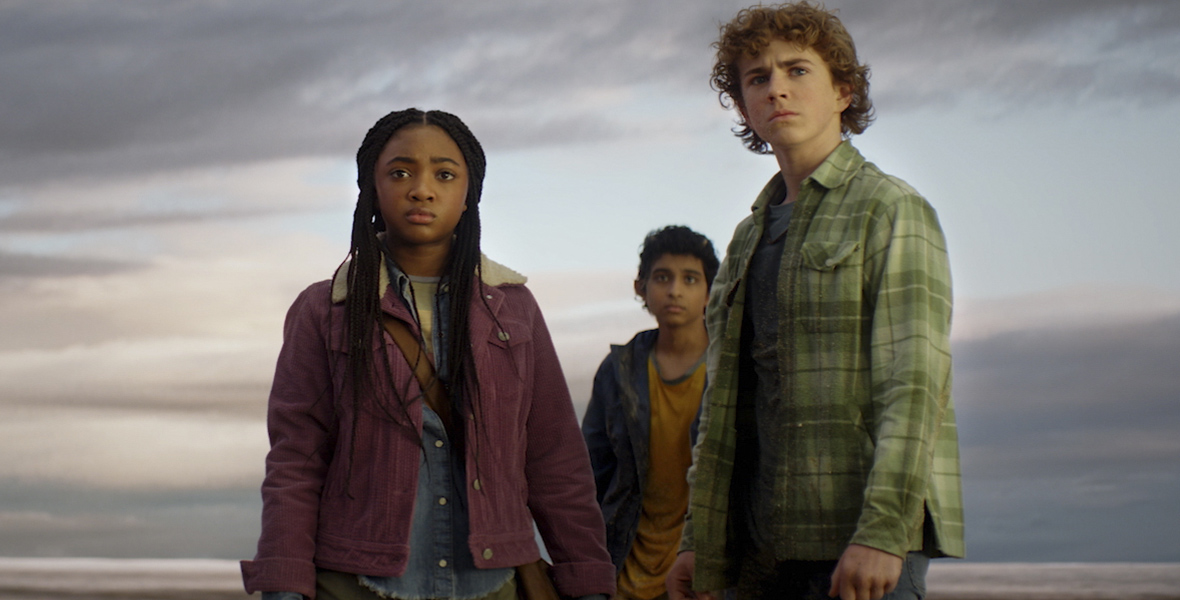 In an image from Disney+’s Percy Jackson and the Olympians, from left to right, Annabeth (Leah Sava Jeffries), Grover (Aryan Simhadri), and Percy (Walker Scobell) are looking concerned at something off camera to the left. The ocean and a vast cloudy sky are seen behind them. Annabeth is wearing a purple jacket and army green pants; Grover is wearing a yellow T-shirt and dark jacket; and Percy is wearing a green plaid button-up shirt and jeans.