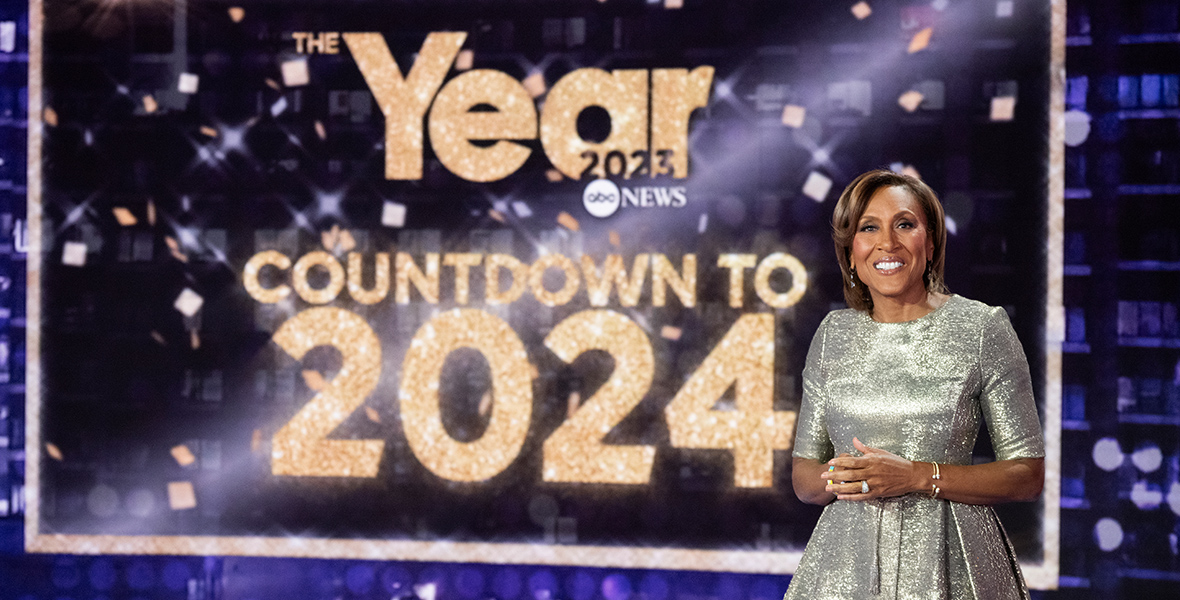 In an image from ABC News’ The Year: Countdown to 2024, host Disney Legend Robin Roberts—wearing a sparkly silver dress—stands in front of a large TV screen displaying the special’s title treatment. She is smiling at the camera.